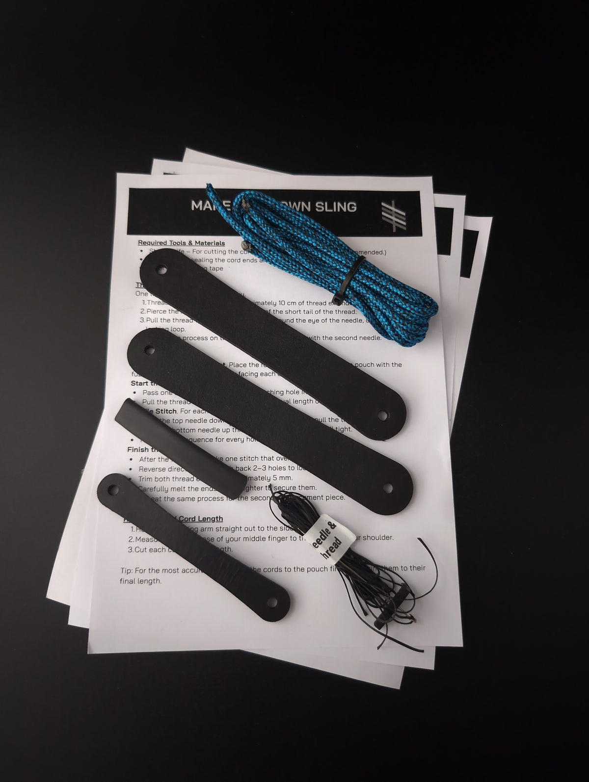 A DIY 'Make Your Own Sling' kit laid out on a black background, featuring four black leather finger loops, a blue braided cord, a small white-and-black instruction card black thread. The main product components are the finger loops and pouchand cord.