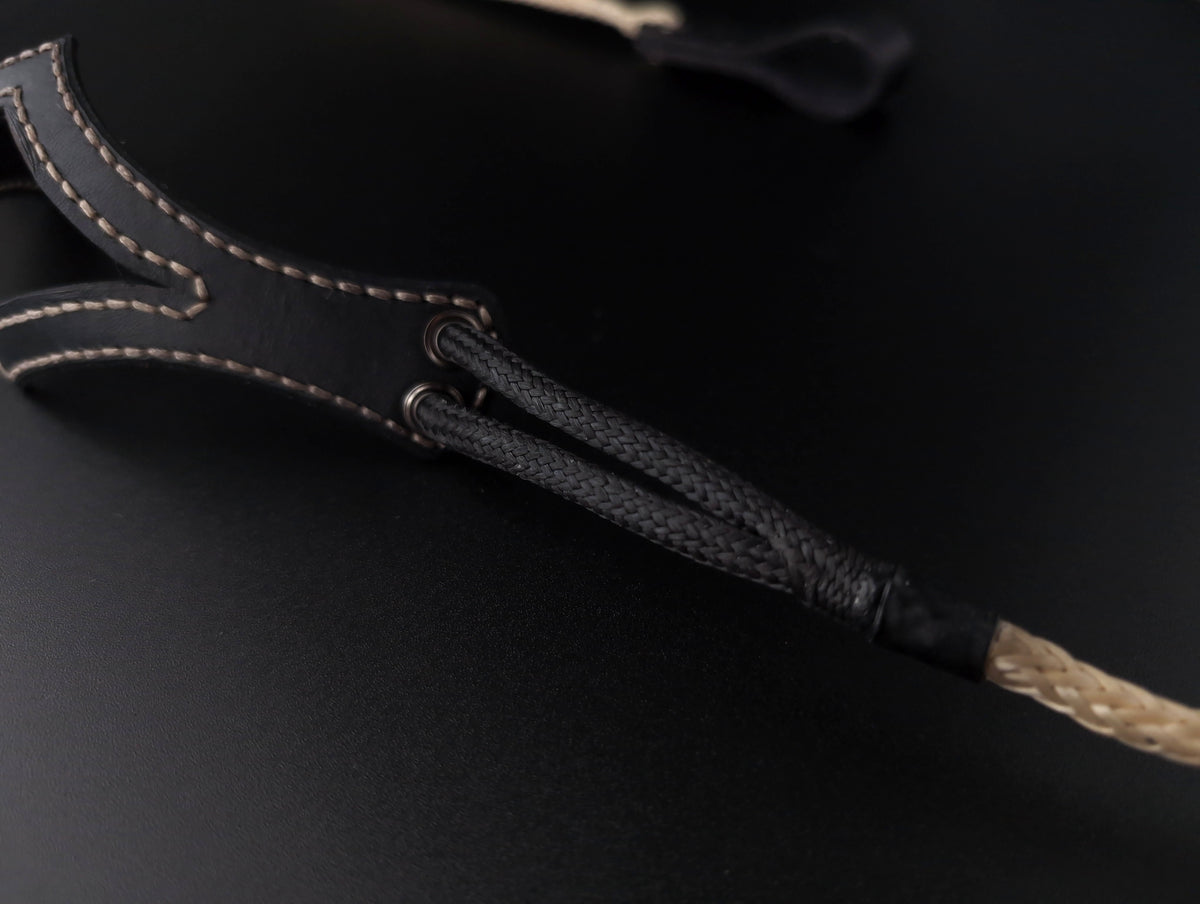 Close-up of a black shepherd sling with light tan contrast stitching and a braided black-and-tan cord, shown on a dark background.