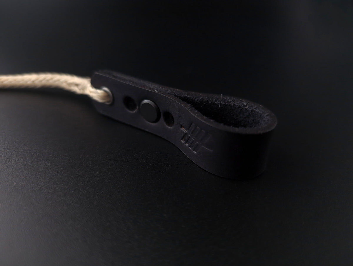 A close-up of a black leather strap with a looped end, featuring a small round button and a light beige rope threaded through a hole, set against a dark black background.