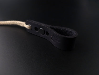 A close-up of a black leather strap with a looped end, featuring a small round button and a light beige rope threaded through a hole, set against a dark black background.
