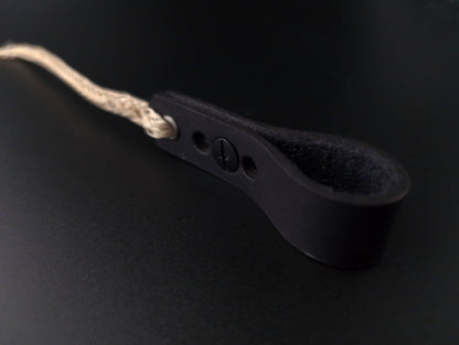 A close-up of a black leather strap with a looped end, a small screw detail, and a beige braided cord attached, set against a dark background.