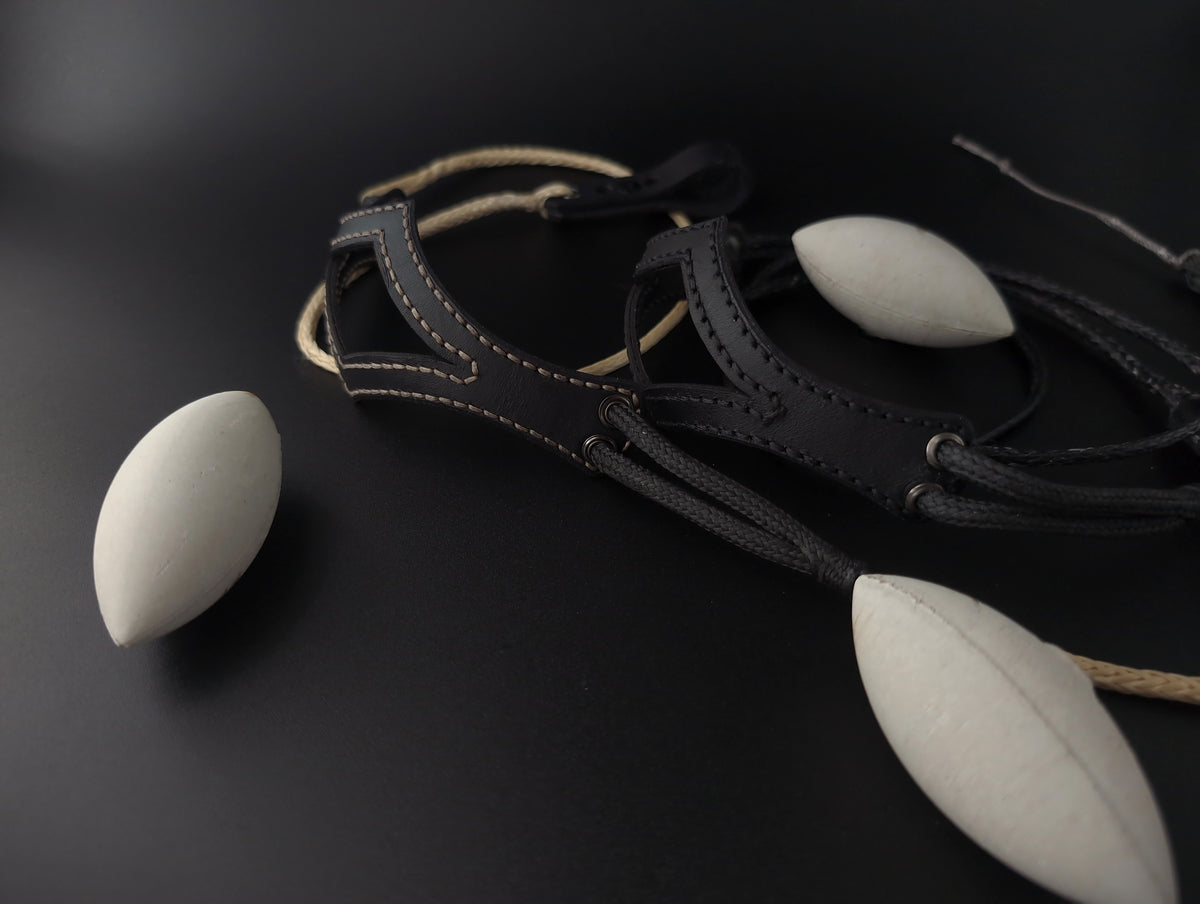 A black leather shephed slingwith light tan and black cord and three aerodynamic concrete sling projectiles, arranged on a dark surface.