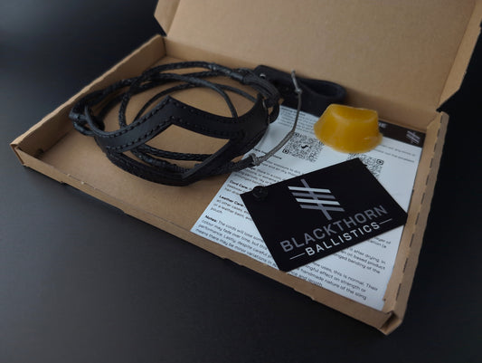 A Blackthorn Ballistics vectran shepherd sling displayed inside its open cardboard packaging on a dark surface. The kit includes a black leather shepher sling with black stitching, a yellow beeswax block, and printed instruction sheets and a black brand tag.