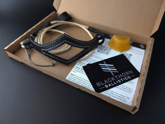A Blackthorn Ballistics vectran shepherd sling displayed inside its open cardboard packaging on a dark surface. The kit includes a black leather shepher sling with beige stitching, a yellow beeswax block, and printed instruction sheets and a black brand tag.